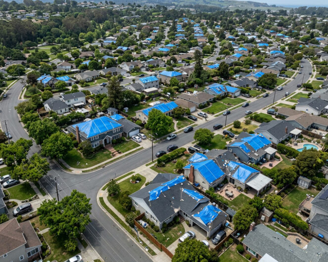 Aerial view of a neighborhood where many homes have blue tarps over storm-damaged roofs